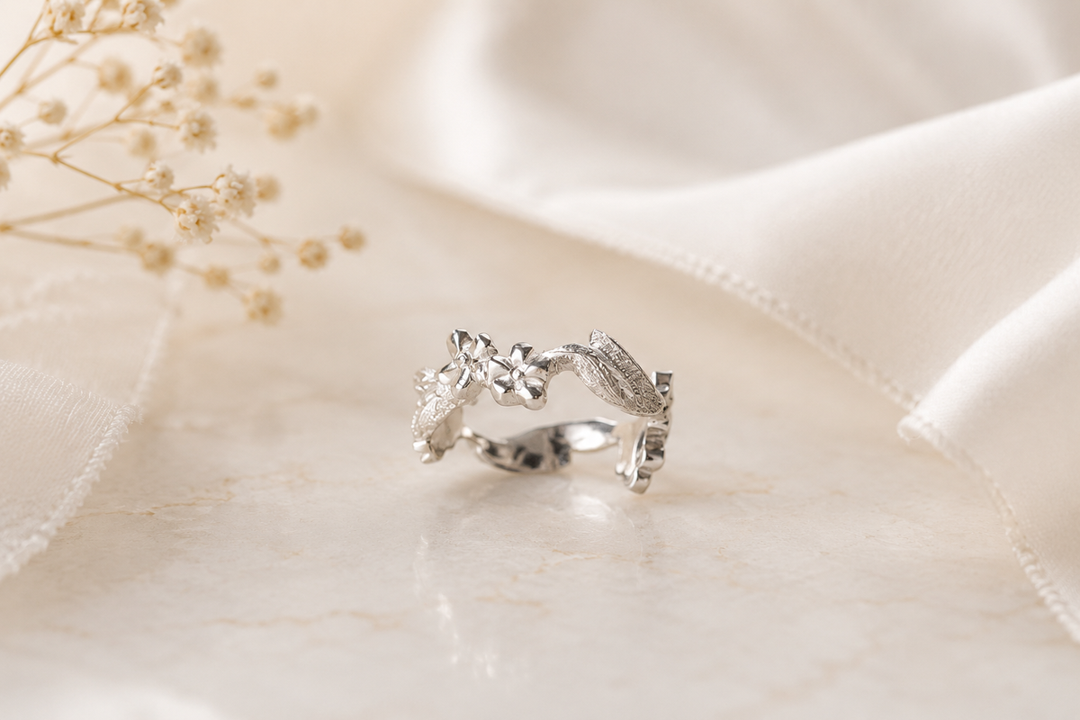 A white gold wedding ring with an engraved, organic leaf-like design, created by Tatiana Marques Jewelry, resting on a light beige marble surface with white satin fabric and dried baby's breath flowers in the background.
