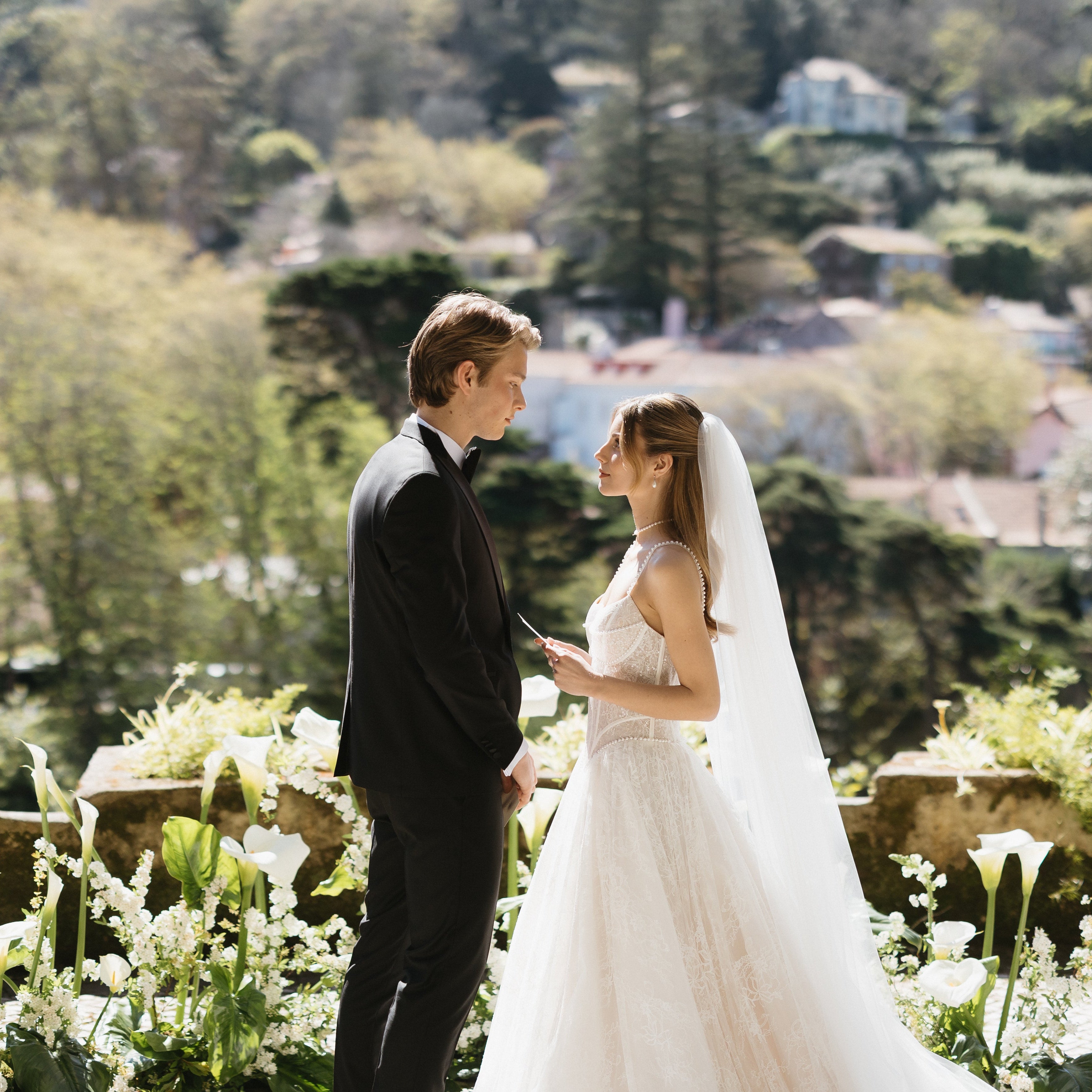 Wedding couple standing in a scenic outdoor setting with greenery and buildings in the background.