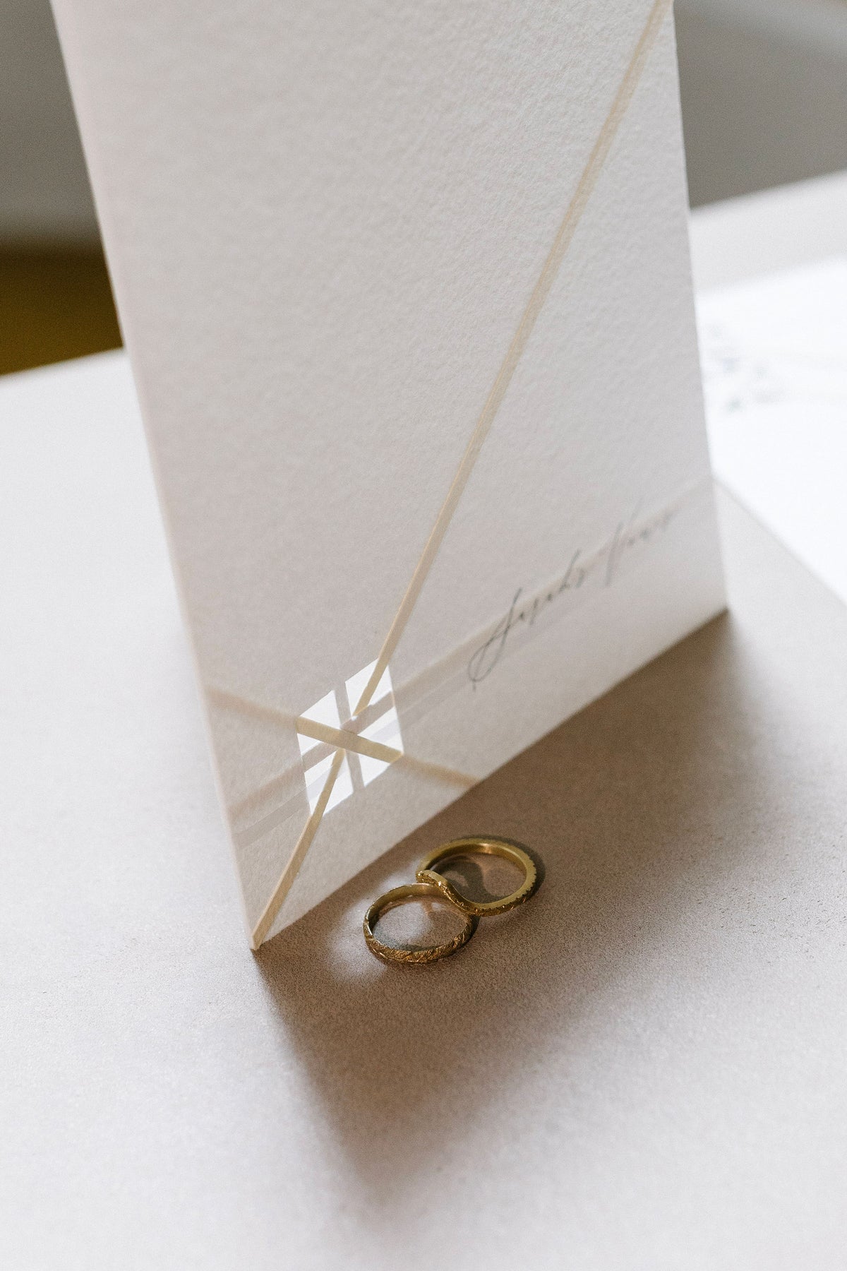 Two gold engraved wedding bands created by Tatiana Marques Jewelry placed beside a tall white invitation card with a geometric cutout design and elegant cursive script.
