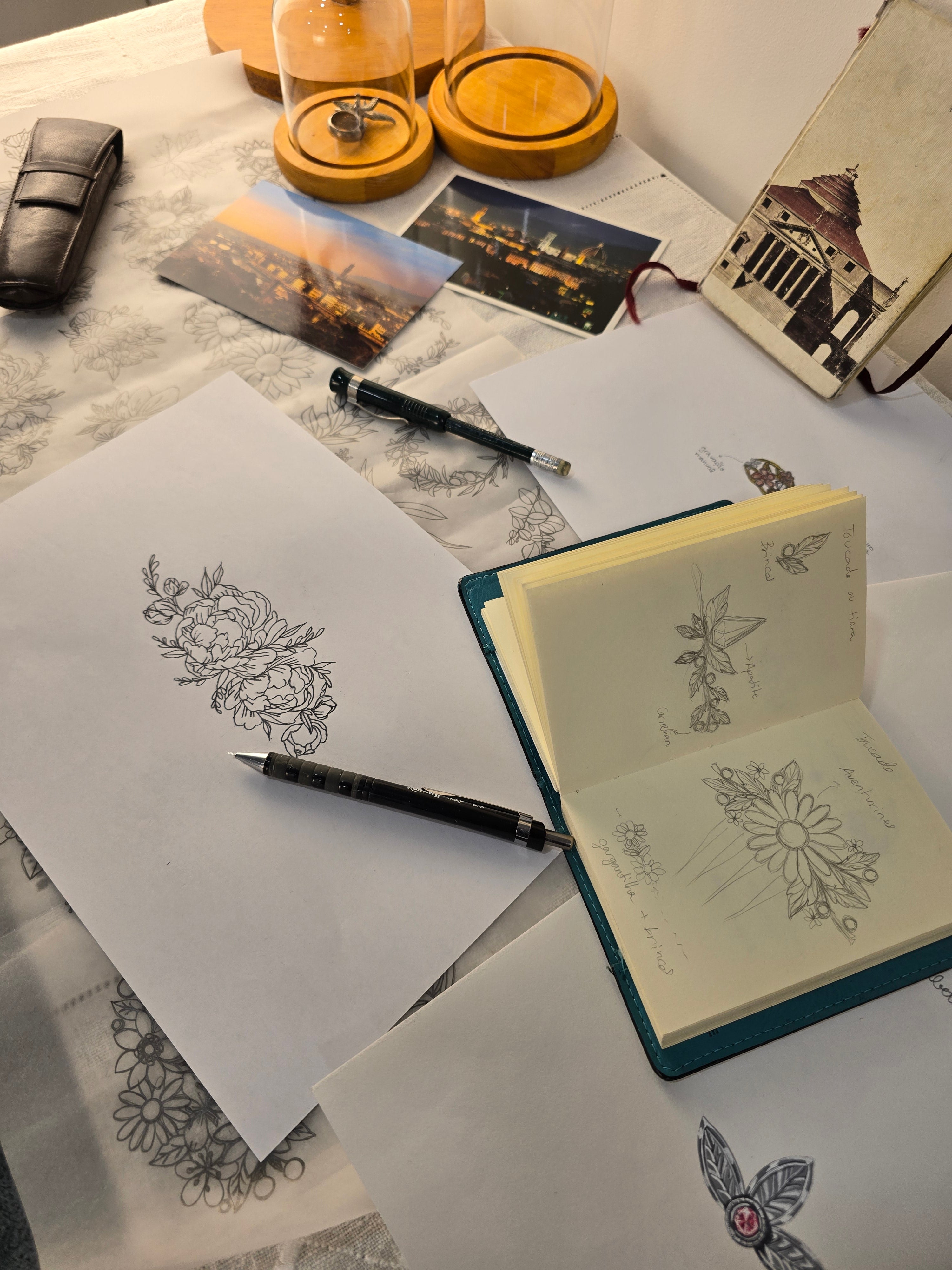 An overhead flat-lay of Tatiana Marques Jewelry's sketchbooks, pens, and small wooden containers on a marble-like surface. An open notebook shows floral and butterfly line drawings, with additional paper sketches and a black pen placed on top.