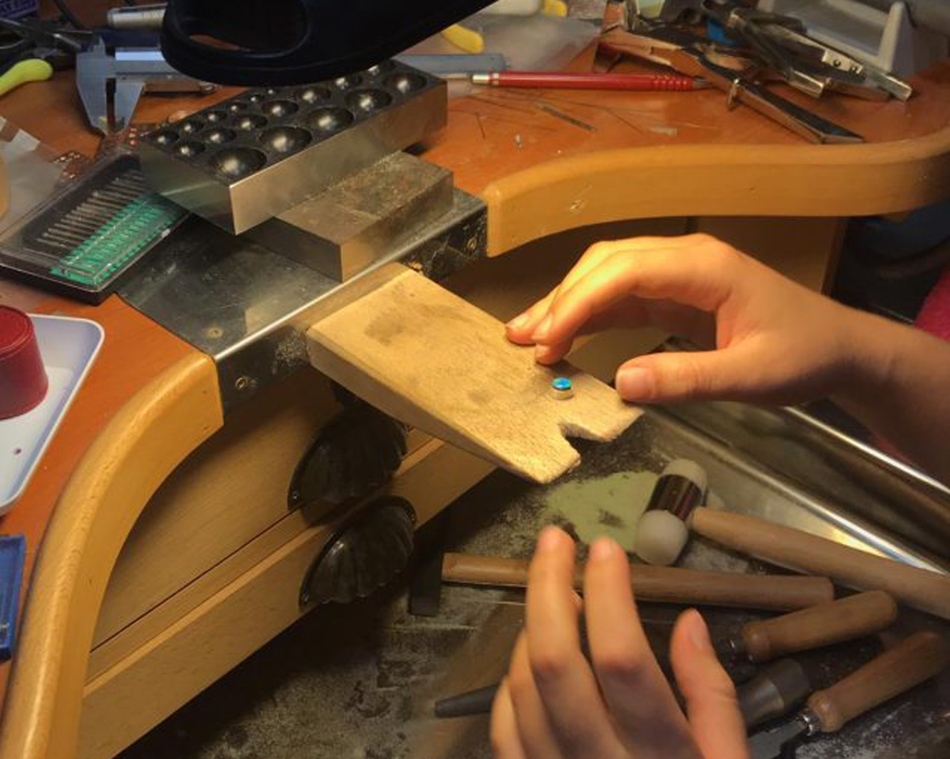 A close-up of the jeweler Tatiana Marques's hands working at a bench, using a small wooden tool to handle a tiny blue gemstone or bead, surrounded by jewelry-making tools and materials.