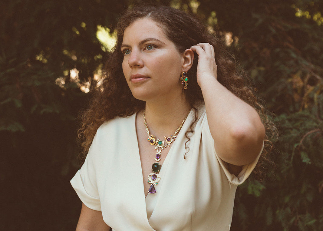 Tatiana Marques with long curly brown hair wearing a cream-colored wrap-style jumpsuit and colorful statement jewelry, Royal Blossom, from Tatiana Marques Jewelry, standing outdoors in front of a dark, sun-dappled forest background.