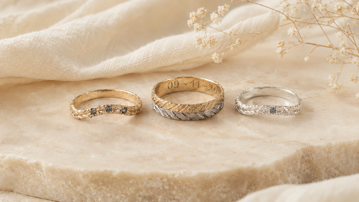 Three hand engraved rings in yellow and white gold, and a two-tone gold ring, created by Tatiana Marques Jewelry, displayed on a beige stone surface with draped cream fabric and dried flowers.