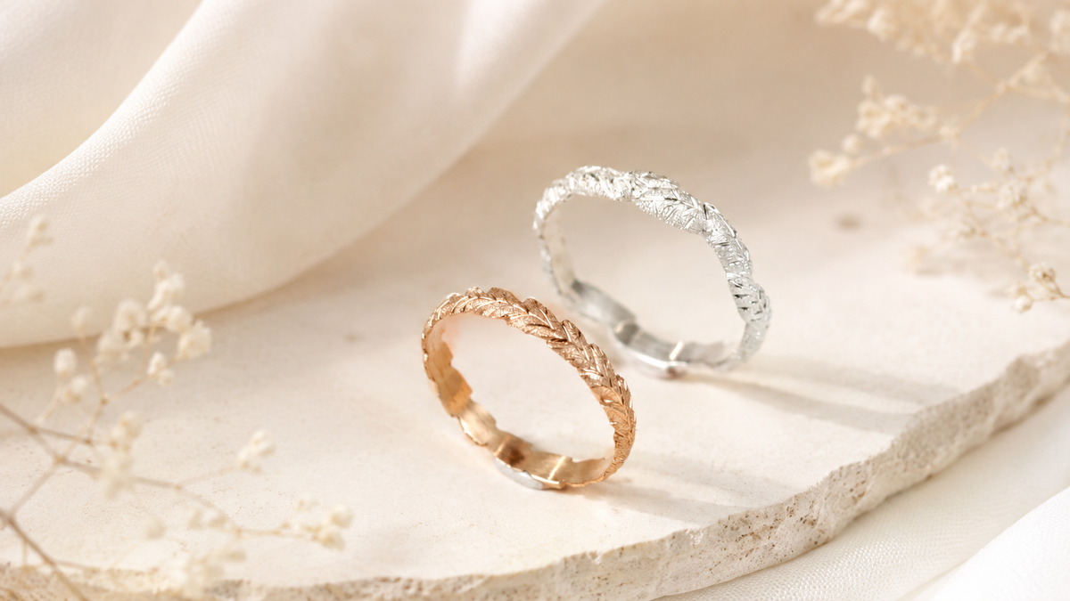 Two engraved wedding band rings, one rose gold and one white gold, created by Tatiana Marques Jewelry and displayed on a light beige stone slab with soft white fabric and dried baby's breath flowers.